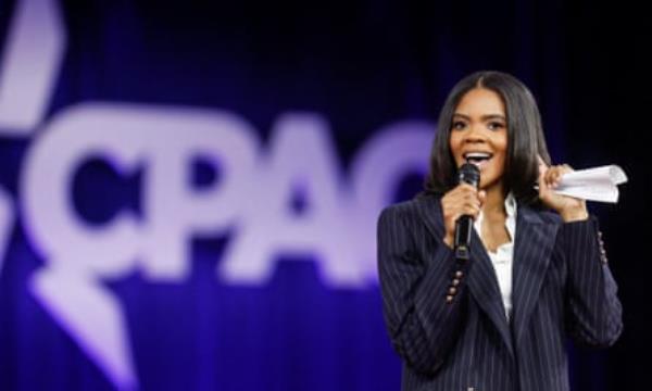 Co<em></em>nservative influencer Candace Owens, wearing a striped suit, talks into a microphone with one hand to her ear and the Co<em></em>nservative Political Action Co<em></em>nference (CPAC) logo is visible in the background. 