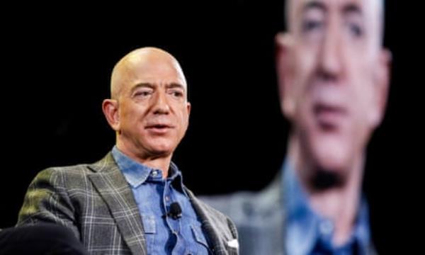 Jeff Bezos, wearing a grey suit with a blue shirt, addresses a co<em></em>nference with a screen with his face on it visible in the background. 