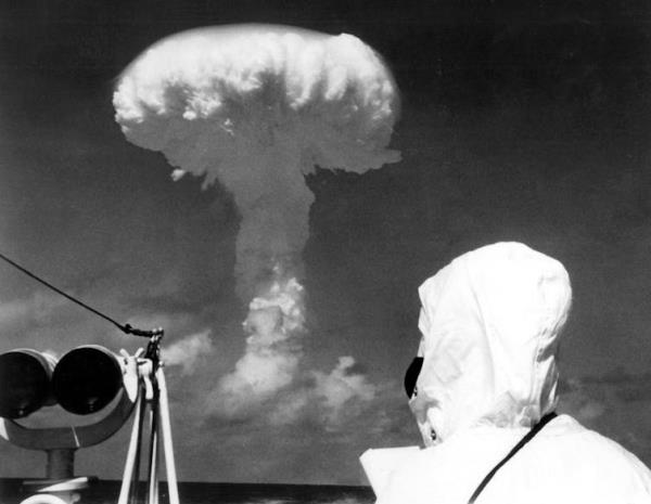 An observer dressed in protective clothing watches a nuclear mushroom cloud.